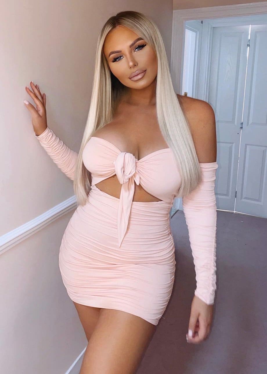 A blonde woman poses with one hand on the wall in a light pink ruched mini dress with a front knot cutout and long sleeves.