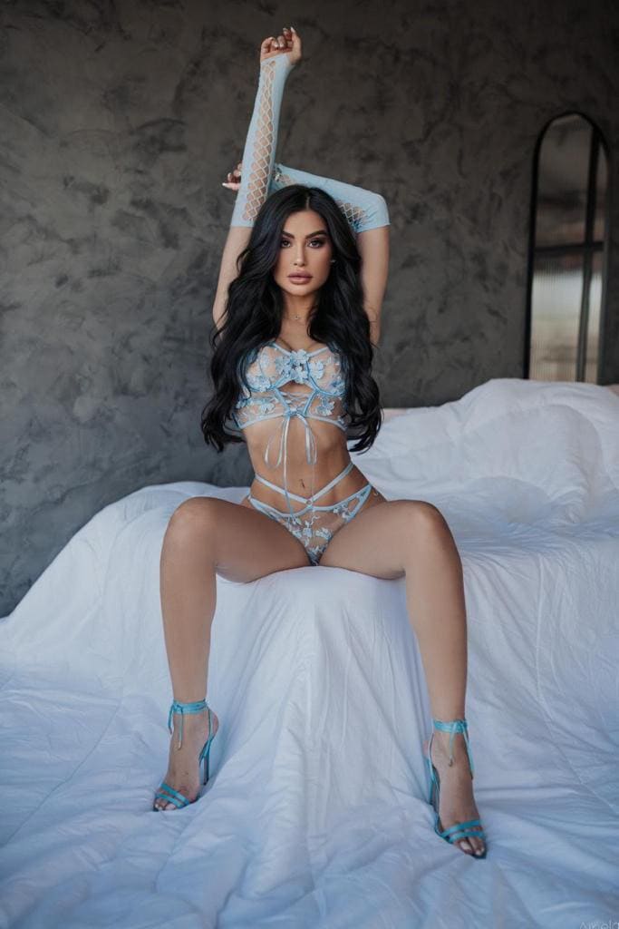 A woman sits on a white bed in light blue lace lingerie, fingerless gloves, and heels, one arm raised against a gray wall.