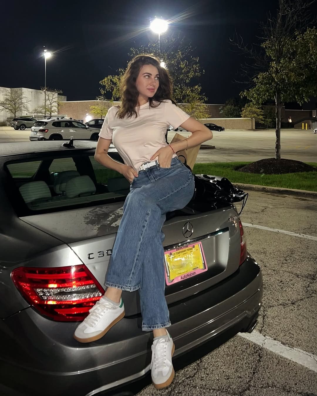 A woman with long wavy brown hair sits on the trunk of a silver sedan in a parking lot, wearing a pink t-shirt, blue jeans, and white sneakers.