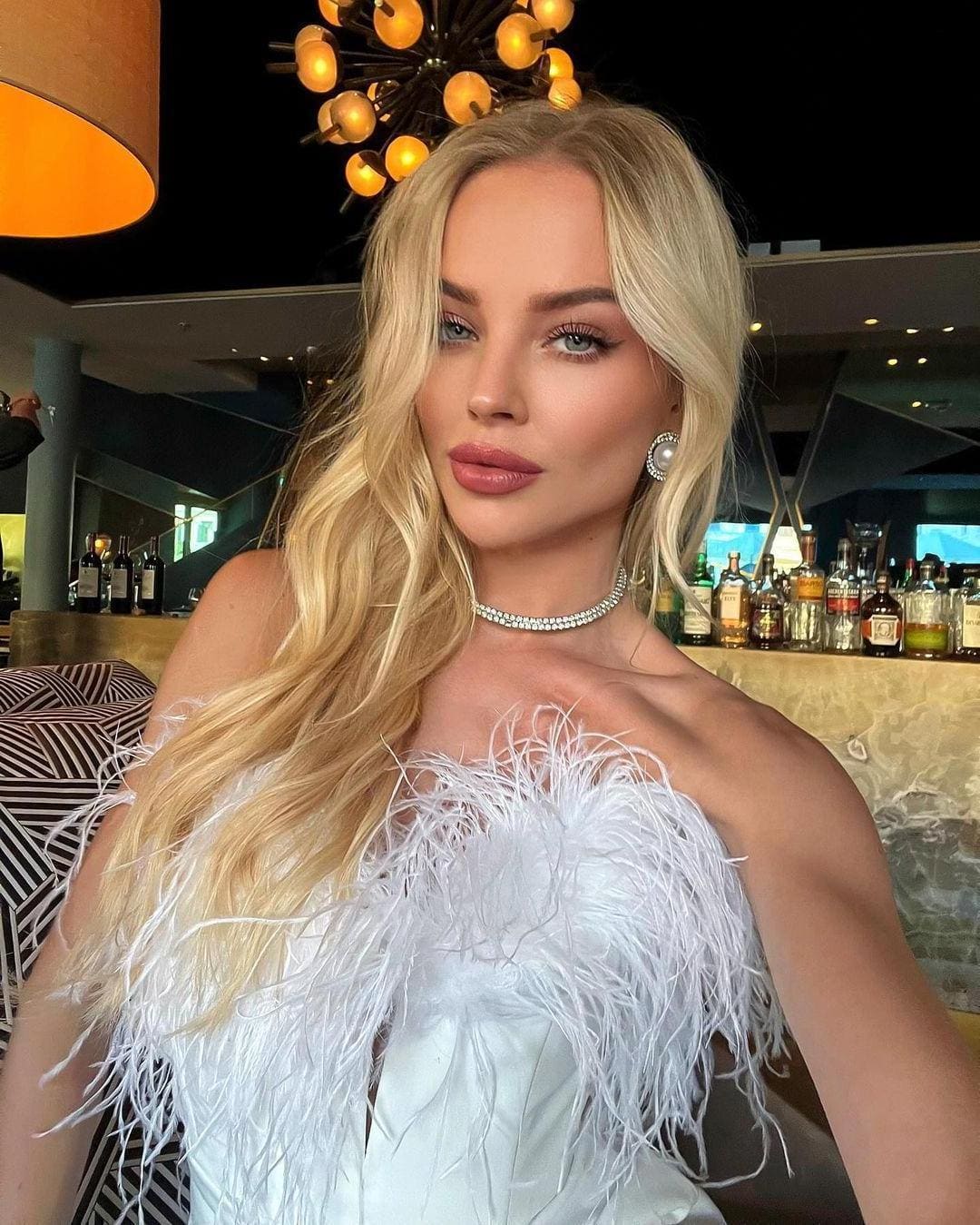 A woman in a white feathered strapless top and beaded choker poses in front of a bar with bottles and a light fixture.