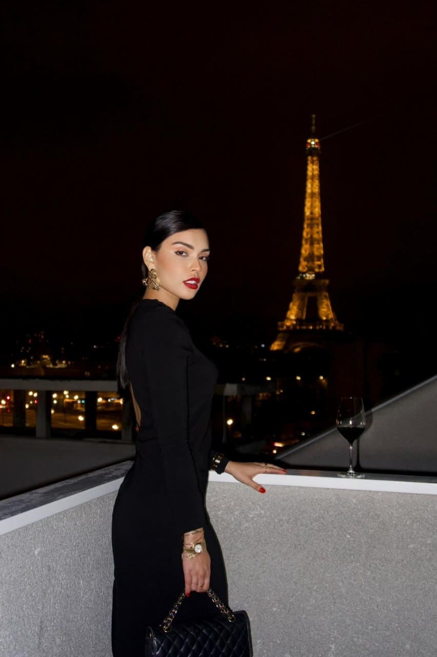 In profile on a night terrace, a woman wears a black backless dress and gold jewelry, holding a handbag near a wine glass and a lit tower.