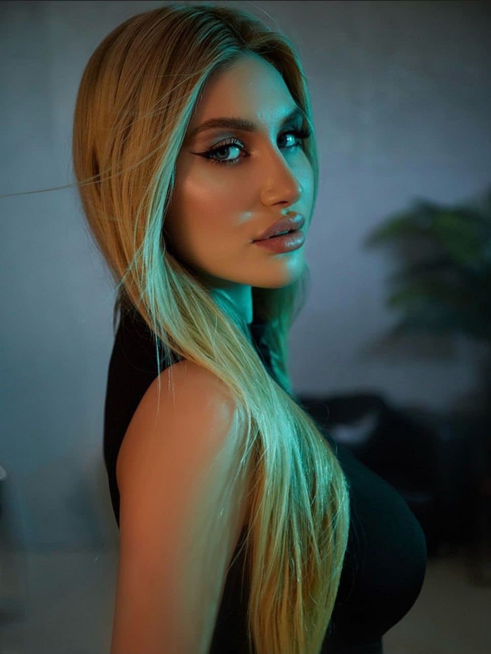 A close-up shows a woman in a black sleeveless top looking over her shoulder in a dim room with a blurred plant behind her.