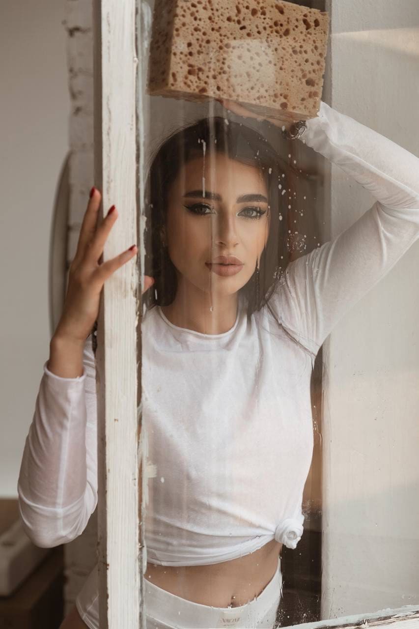 A woman in white crop top and bottoms stands behind glass, holding a sponge overhead as water drips down the pane.