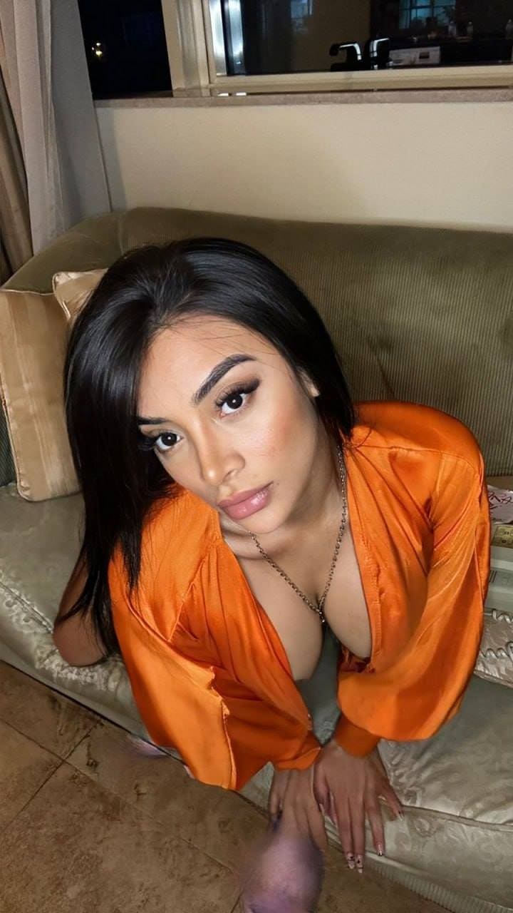 Kenny sits on a beige couch in an open orange satin shirt over a light top, with a window and curtains behind her.