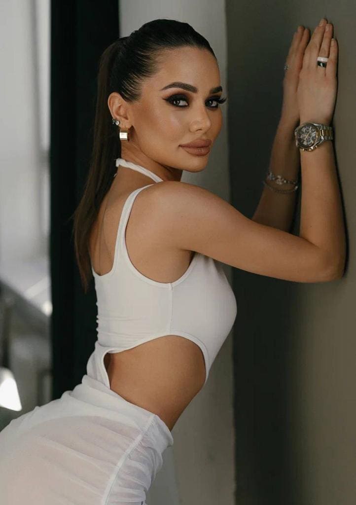 With a high ponytail, she leans against a wall in a white cut-out halter top and sheer skirt, hands pressed flat, looking over her shoulder.
