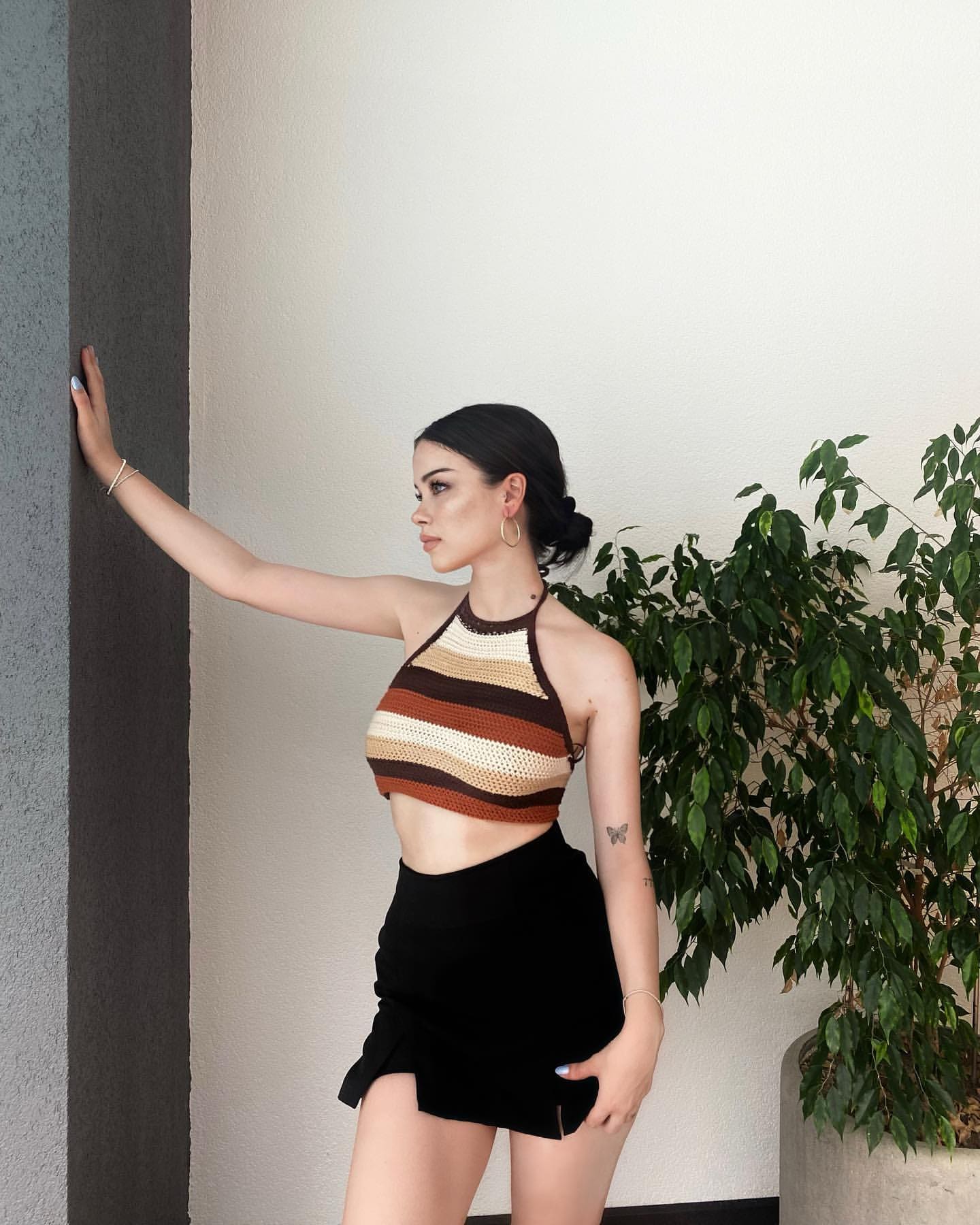A woman in a striped halter top and black mini skirt stands in profile against a white wall, hand on a gray pillar.