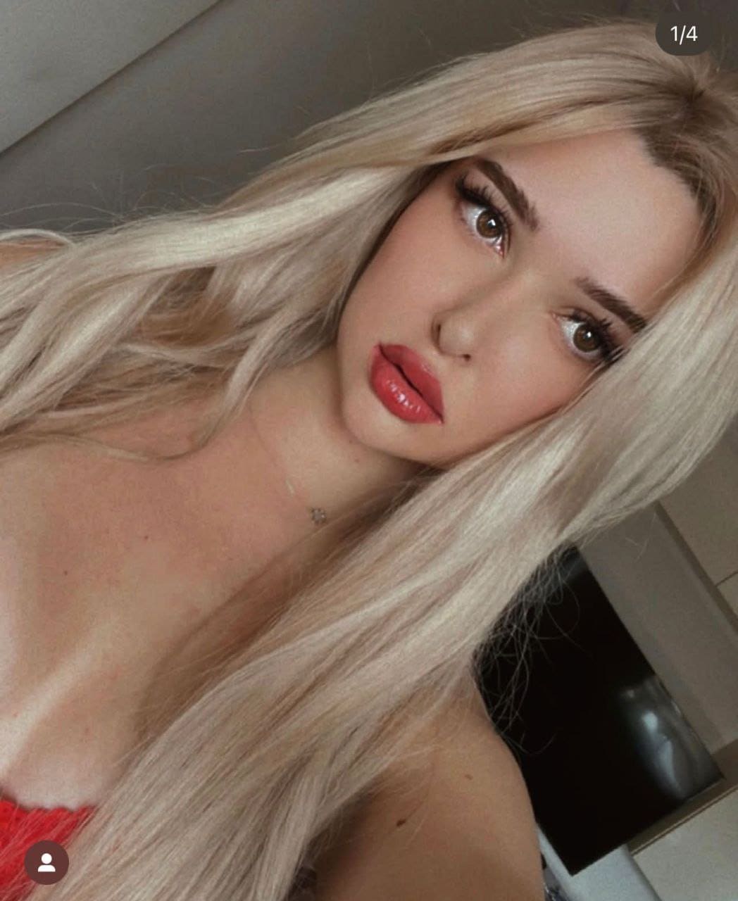 A close-up selfie of a blonde woman in a red strapless top and necklace, posed indoors against a dark screen background.