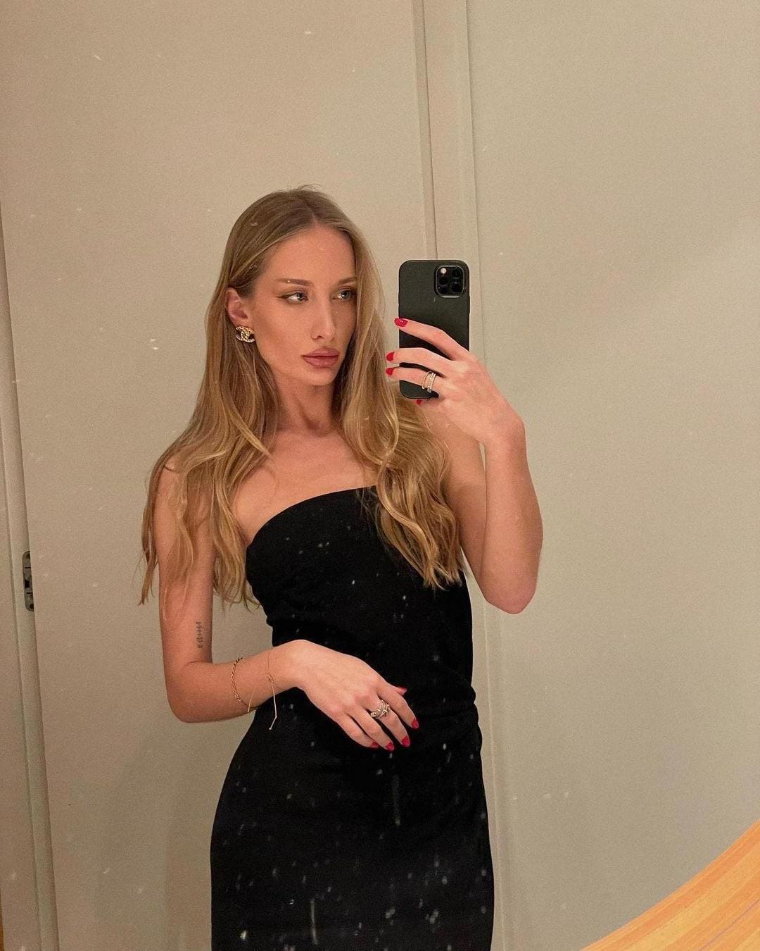 A mirror selfie shows a woman in a black strapless dress holding a dark smartphone, her other hand resting on her hip against a plain light wall.