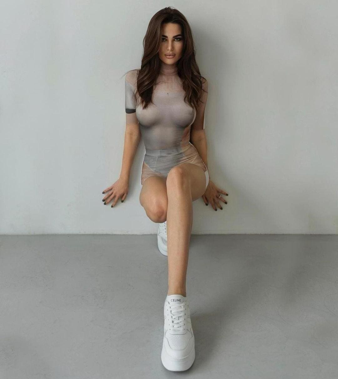 A woman sits on a gray floor against a white wall, wearing a sheer light-colored short dress with a high neckline and white sneakers.