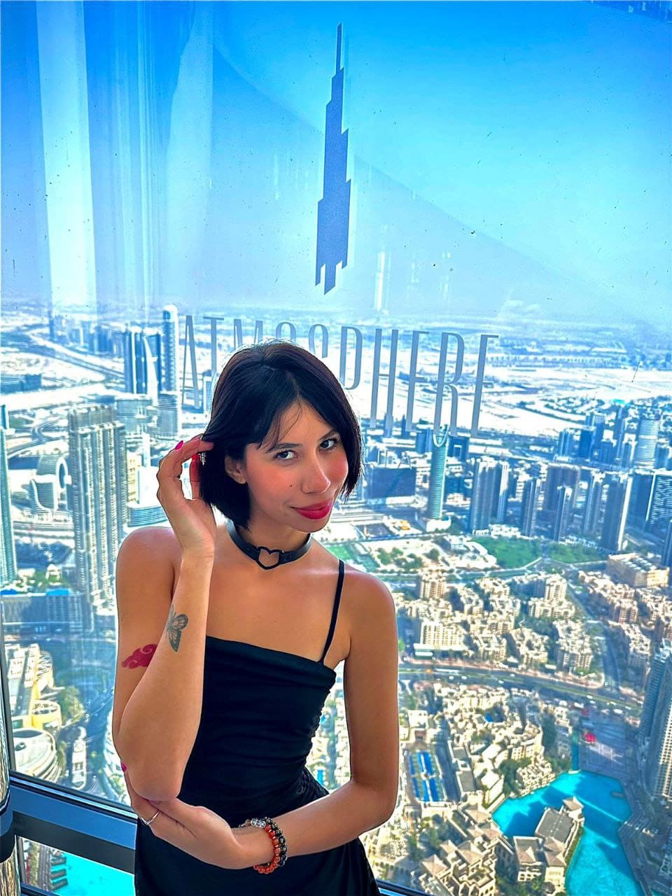 In a black spaghetti-strap dress and choker, she stands by a window with a city view, posing with one hand near her head.