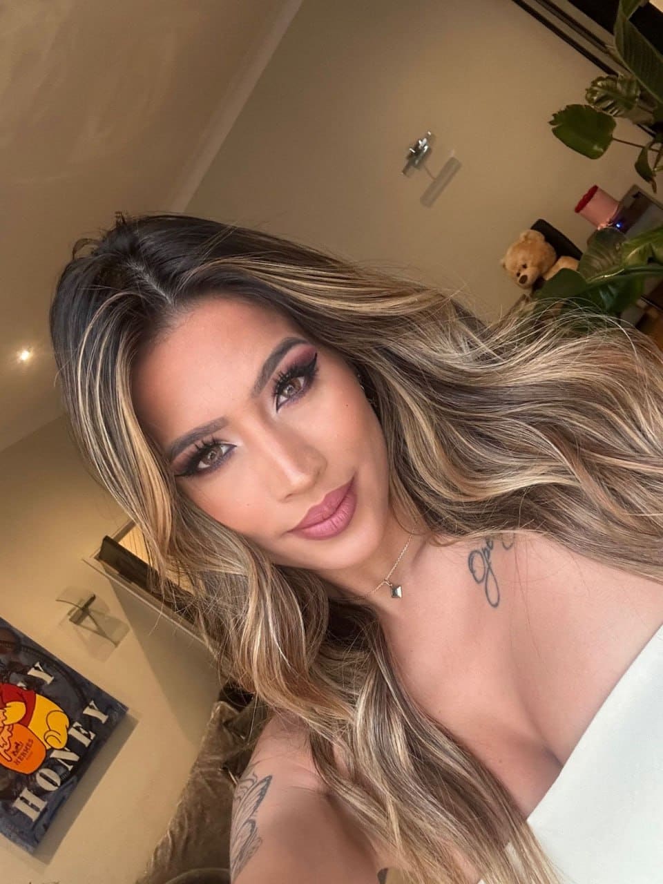 Close-up selfie of a woman with blonde-highlighted hair in a white off-the-shoulder top, showing arm tattoos, with a plant and poster behind.