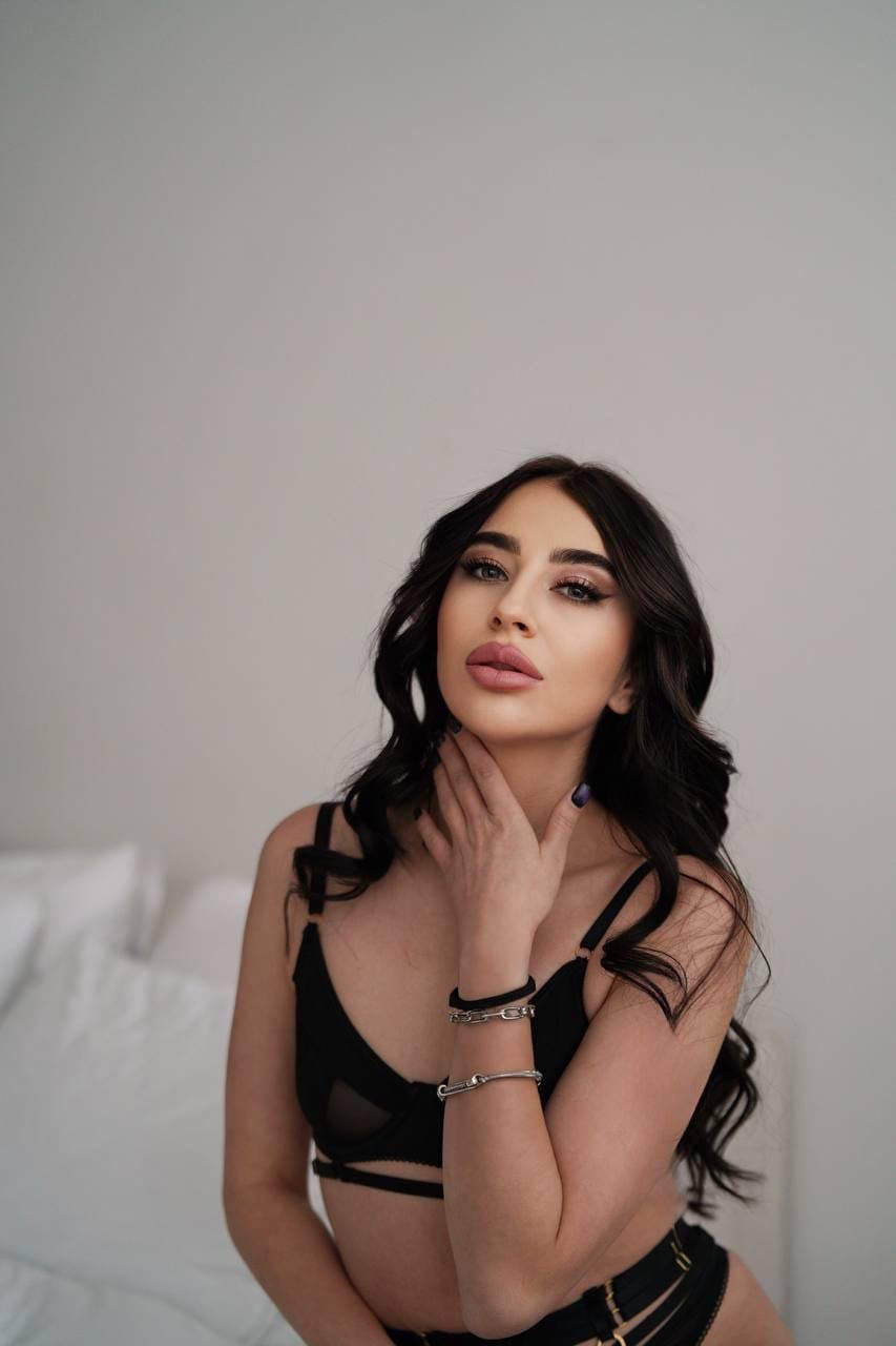Eira sits before a white bed in black strappy lingerie, resting a hand on her neck, with bracelets on her wrist.