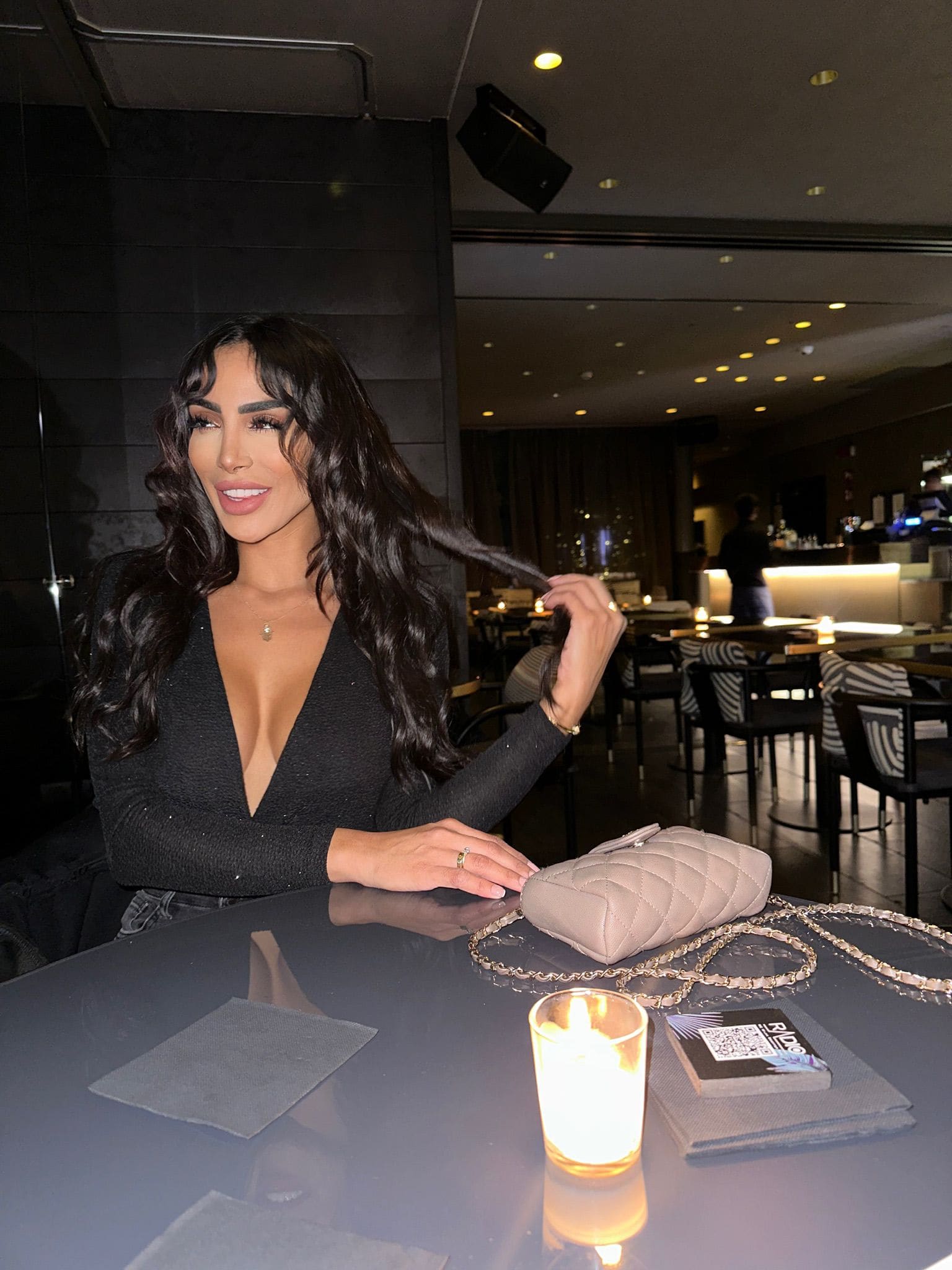 Seated in a dim restaurant, she wears a black V-neck top, touching her hair near a beige handbag and lit candle.