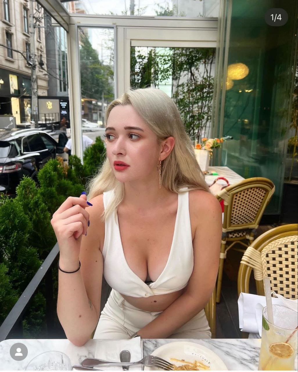 ANTONIA sits at a marble table in a glass-walled room, wearing a white sleeveless top, with food and lemonade in front of her.