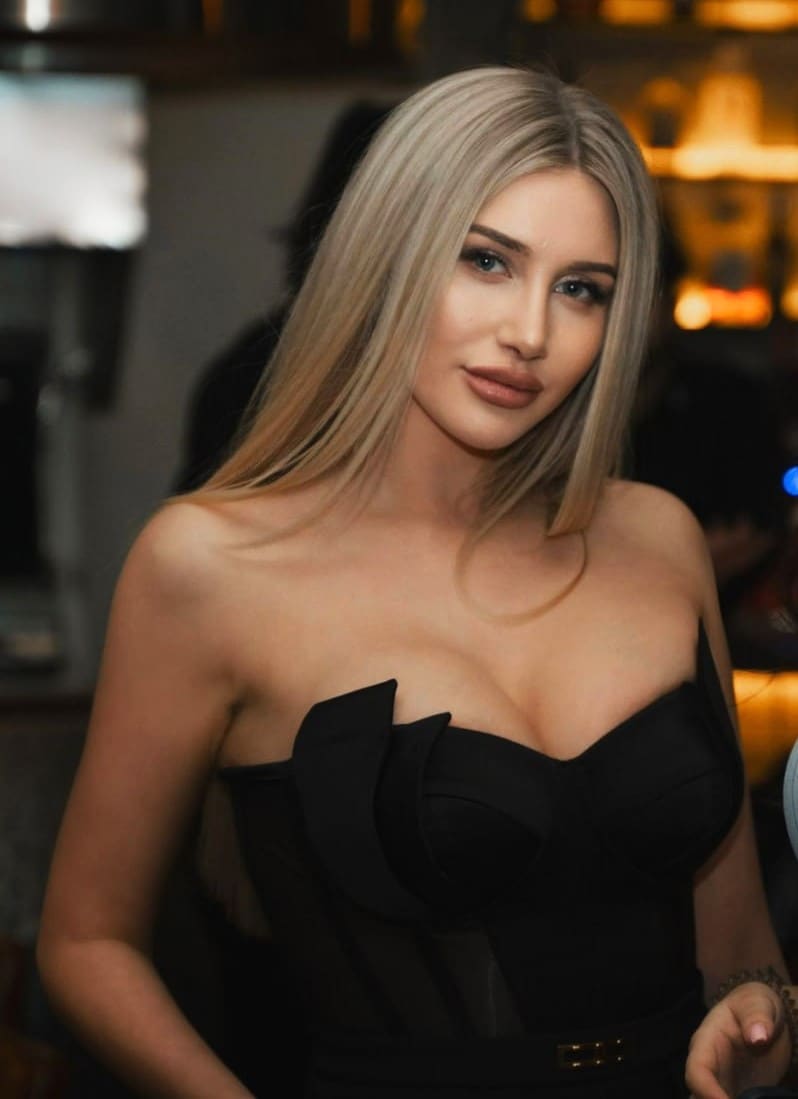 A woman in a black strapless dress with a ruffled neckline and gold-buckled belt stands in a dimly lit room with warm lights.