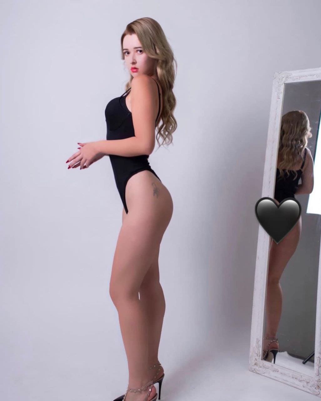 A blonde woman in a black bodysuit and heels stands in profile against a white wall, her back visible in a framed mirror.