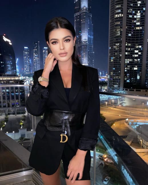 Celestine stands on a rooftop at night, wearing a black blazer, shorts, and a wide belt, with a city skyline behind her.