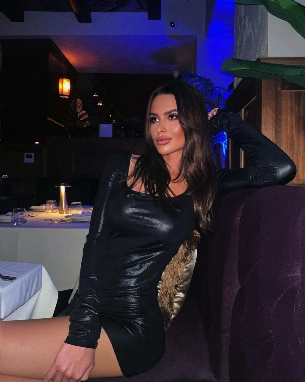 A woman sits on a purple couch in a dim restaurant, wearing a tight black long-sleeved mini dress with one arm raised behind her head and legs crossed.