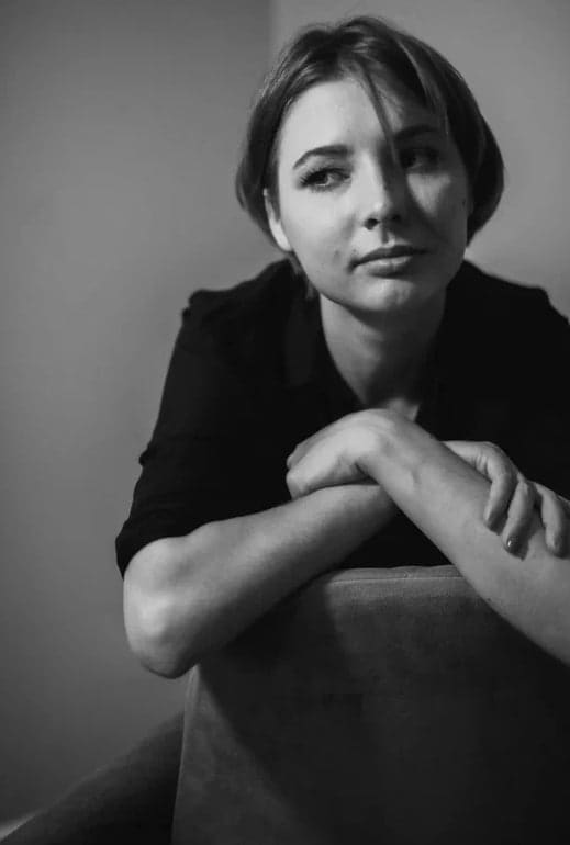 Margo leans forward with crossed arms on a chair back, wearing a dark collared shirt in this black-and-white portrait.