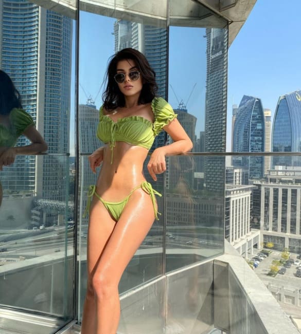 A woman in a lime-green bikini and sunglasses stands on a glass balcony, framed by tall city skyscrapers under blue sky.