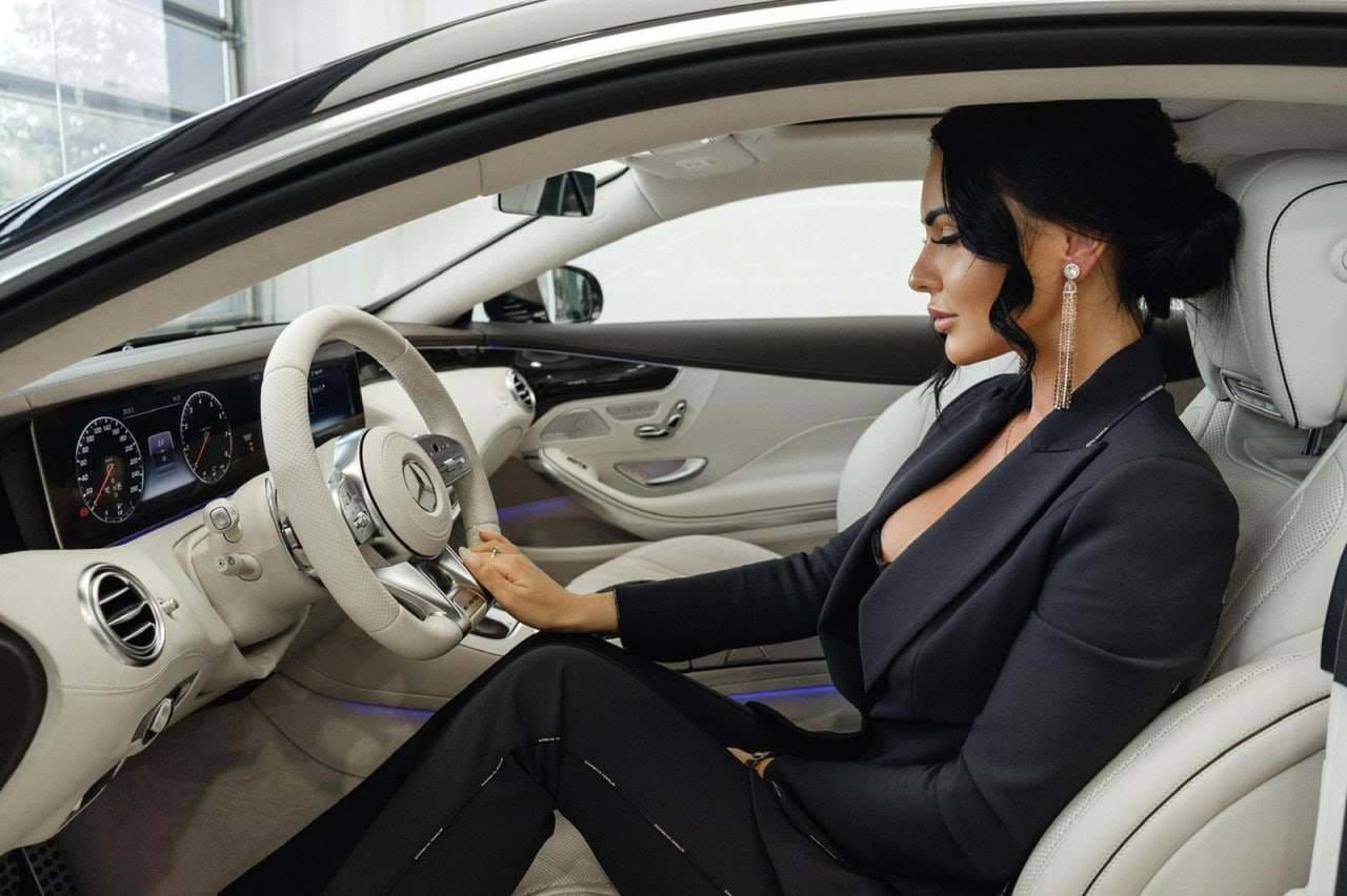 A woman with a low bun sits in a car driver's seat, wearing a black blazer and trousers, hand on the steering wheel.