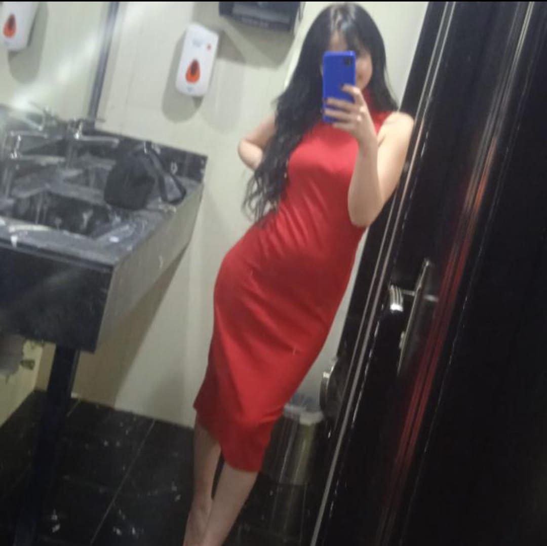 A woman in a red sleeveless dress takes a bathroom mirror selfie, with sink and dispensers visible behind her.