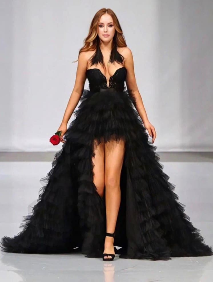 A woman in a strapless black gown with a tulle skirt and slit holds a red rose on a light runway.