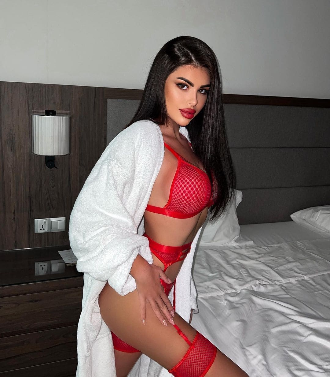 A woman stands in a bedroom in red mesh lingerie and an open white bathrobe, posing with one hand on her hip beside a bed.