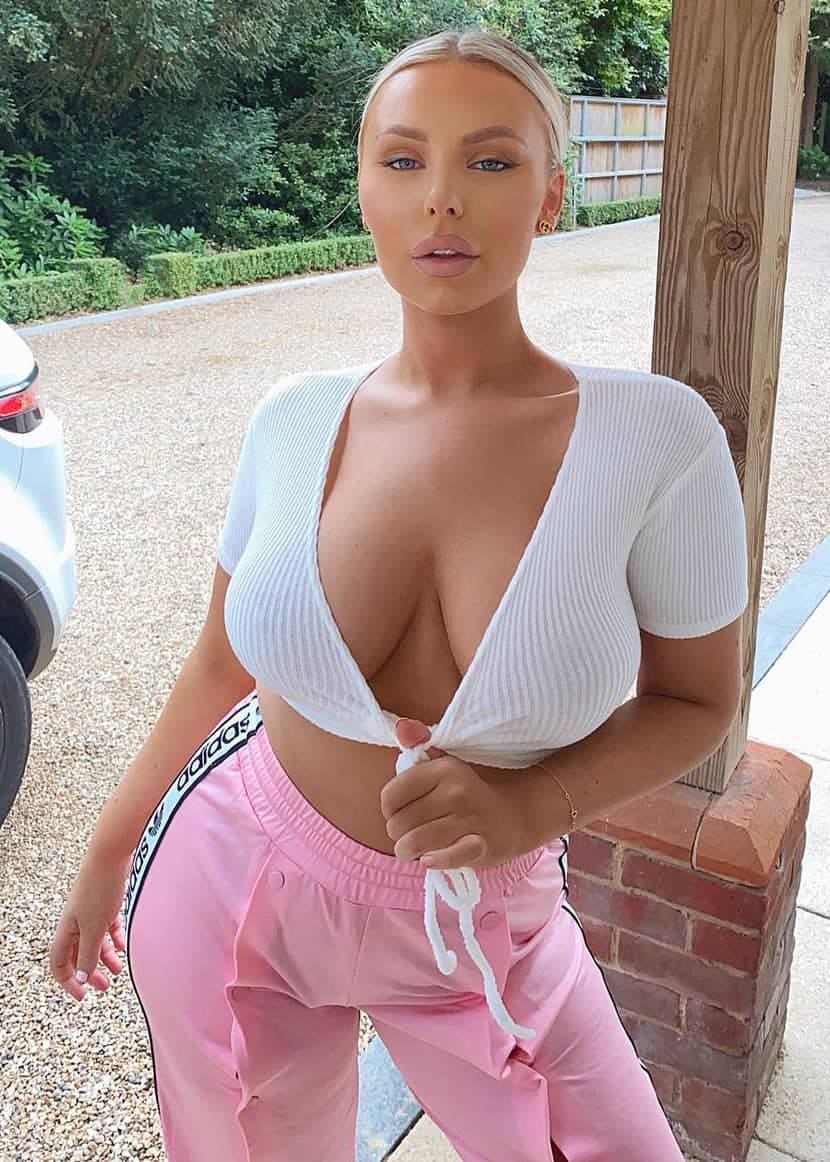 A blonde woman leans against a brick pillar outdoors, wearing a tied white top and pink track pants on a gravel driveway.