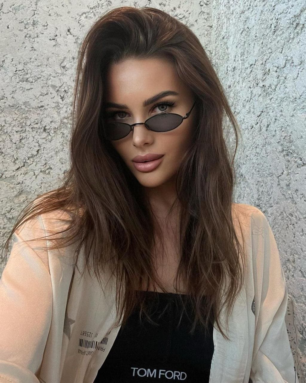 A woman with wavy hair wears oval sunglasses and an open light shirt over a black top, standing against a rough white wall.