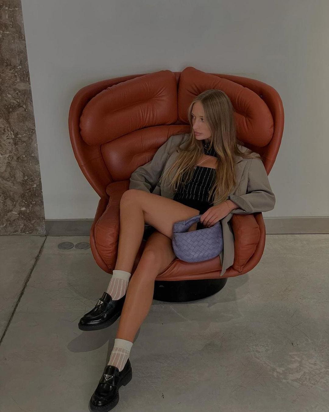 A woman sits in a rust-colored leather armchair wearing a grey blazer, black top, white socks, and loafers, holding a purple handbag on her lap.