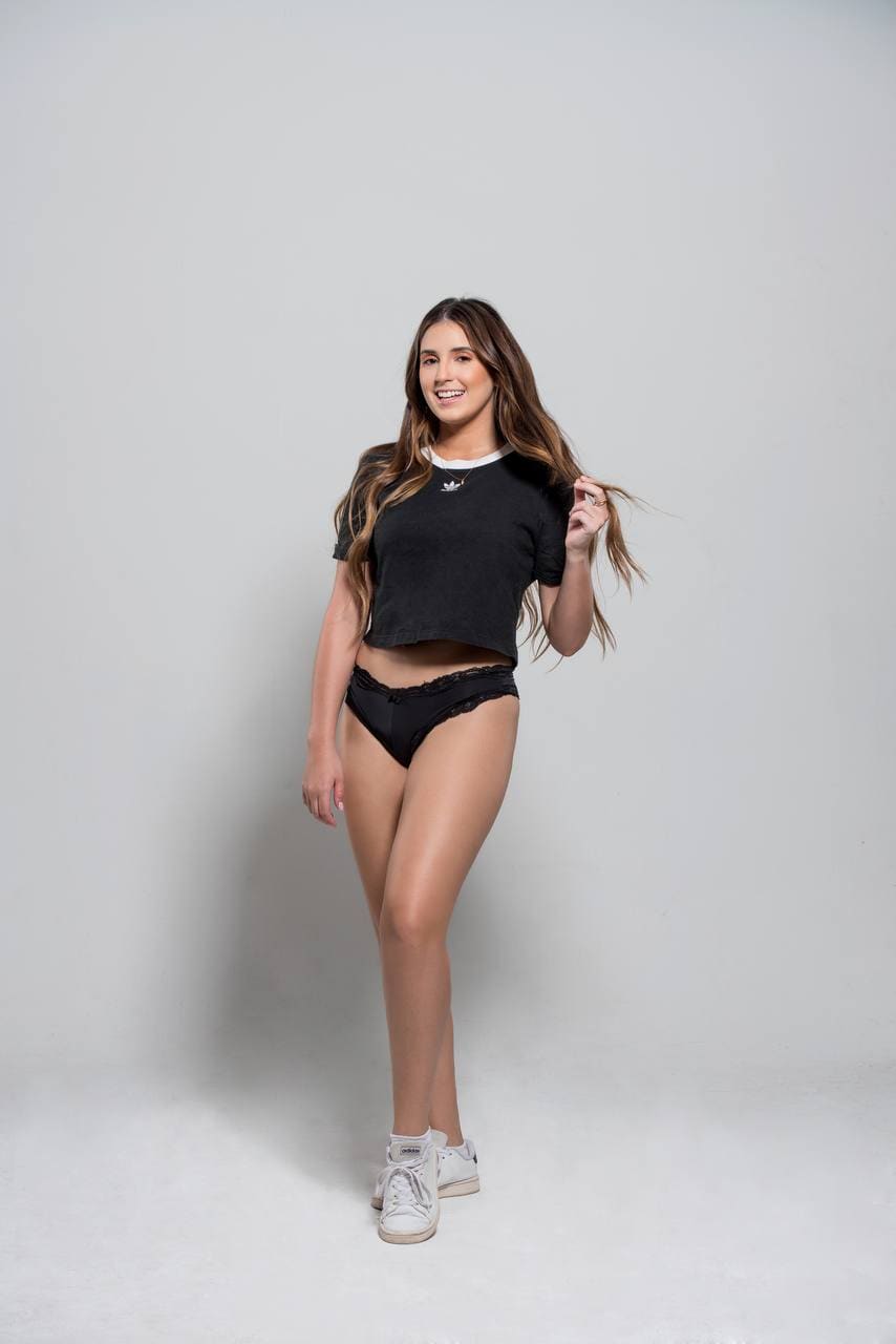 A woman stands in a studio wearing a black t-shirt, lace underwear, and white sneakers, lifting a strand of hair.