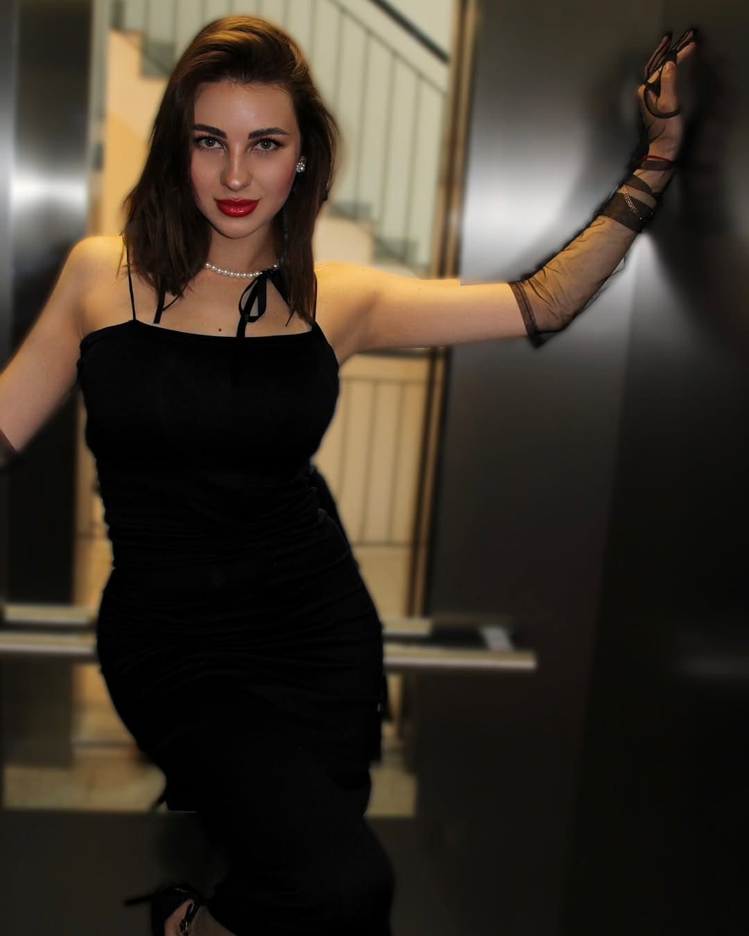 Penelope stands in an indoor stairwell wearing a black spaghetti-strap dress, pearl choker, and sheer black arm sleeves.