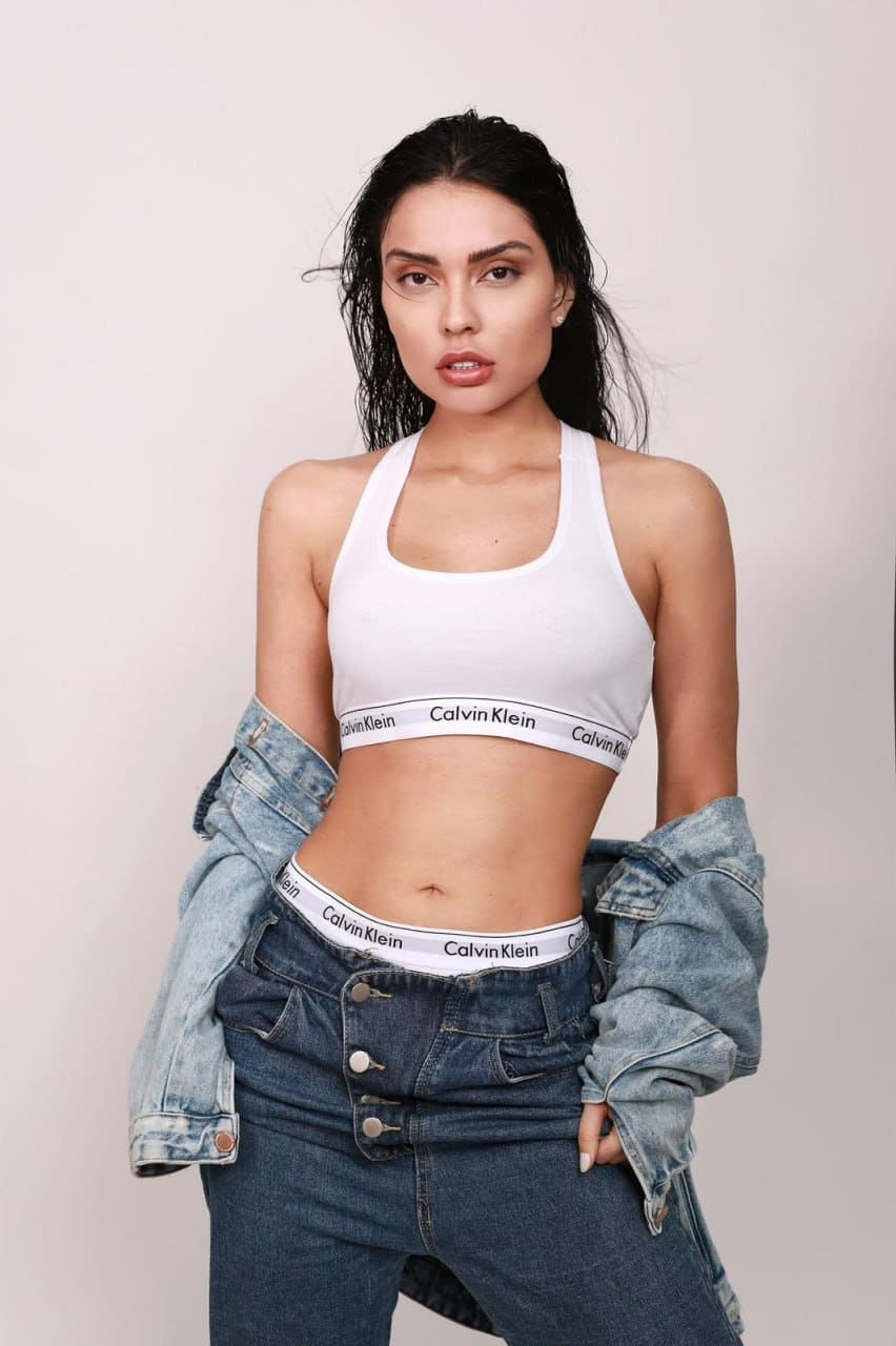 Messika stands against a light background with wet dark hair, wearing a white sports bra, low-rise jeans, and a denim jacket off her shoulders.