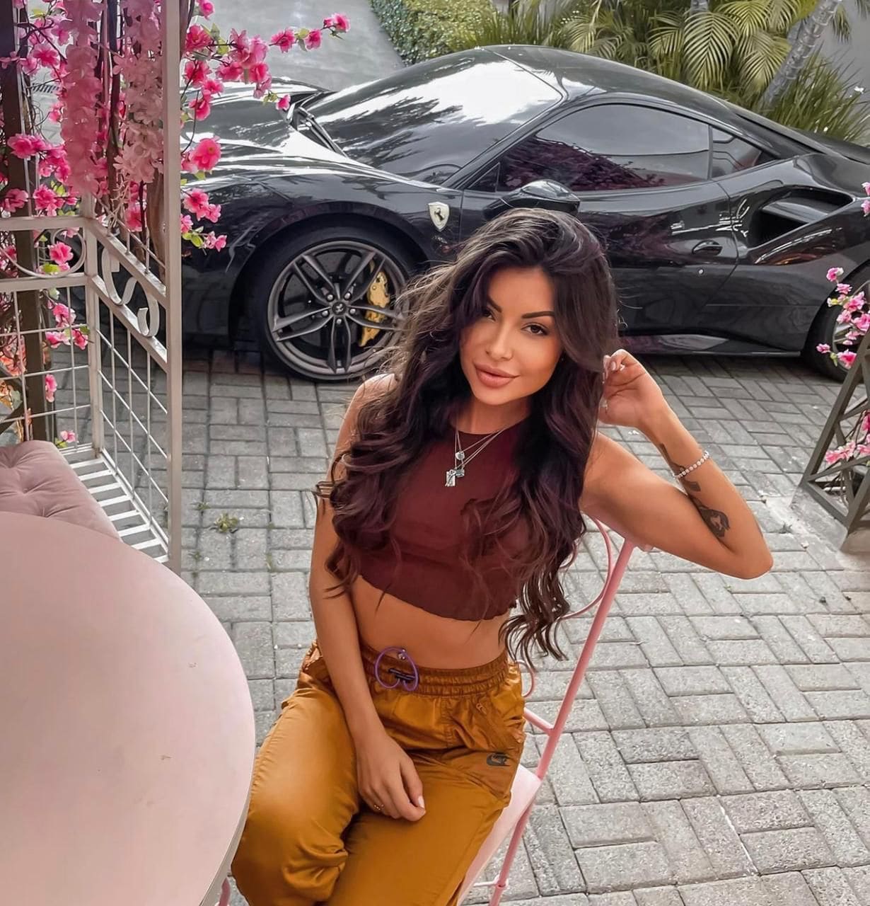 Nina sits on a pink chair in a maroon cropped top and mustard pants, with a black sports car and pink flowers behind her.