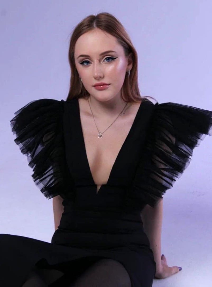 A seated woman in a black dress with ruffled tulle sleeves poses against a plain lavender backdrop.