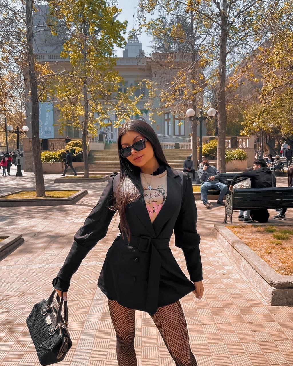A woman stands on a paved plaza in a black blazer dress, fishnets, and sunglasses, holding a handbag near a large building.