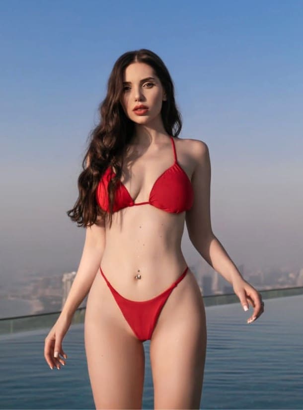 A woman in a red two-piece bikini stands by a pool, with a hazy city skyline and blue sky behind her.