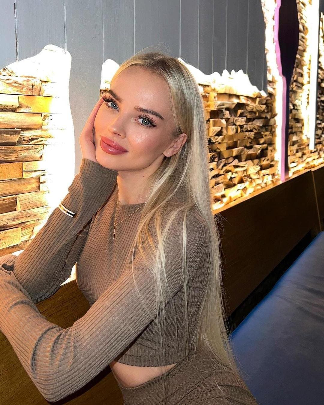 Zoelle rests her chin on her hand in a brown ribbed crop top, seated before stacked firewood with warm backlighting.