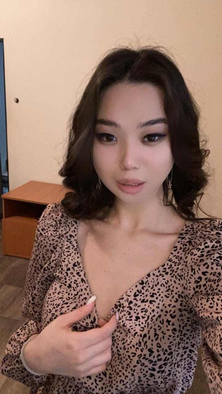 A woman takes an indoor selfie wearing a leopard-print top with a deep V-neck, holding the neckline, with a wooden cabinet and plain wall behind.