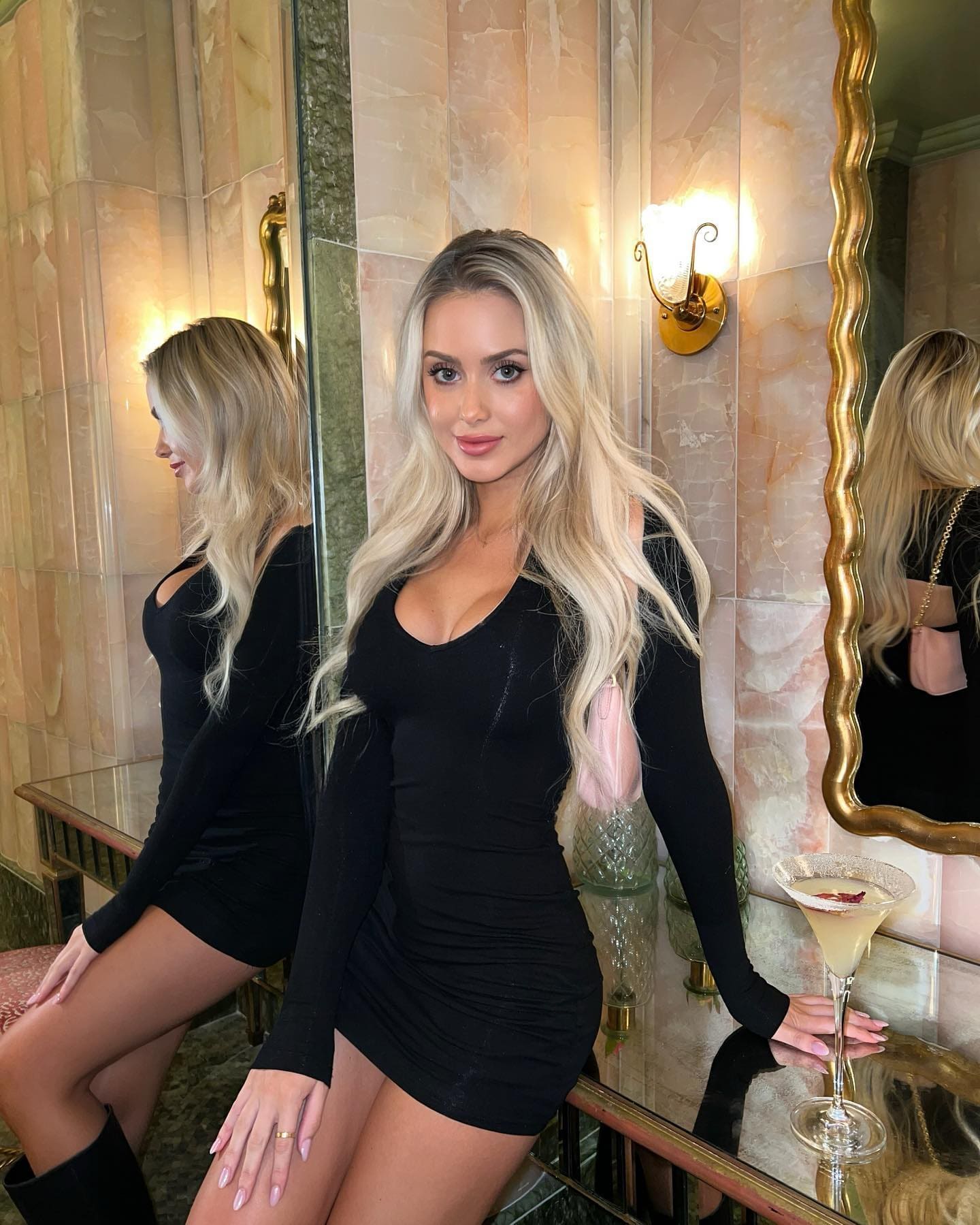 A blonde woman in a tight black long-sleeved mini dress holds a martini glass in front of a marble wall and mirror.