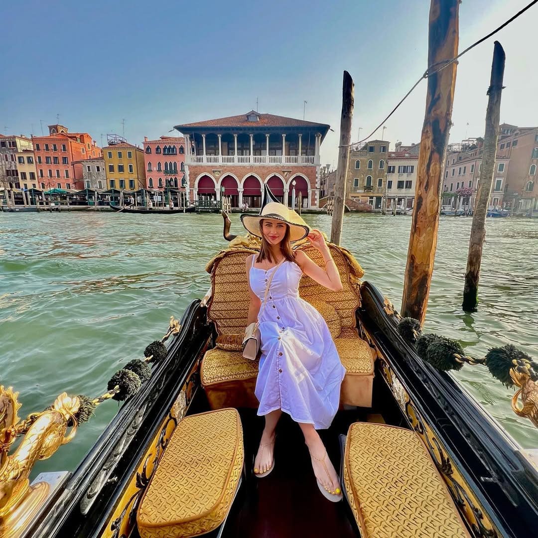 A woman with long brown hair sits in an ornate boat on a canal, wearing a light blue sleeveless dress and a wide-brimmed straw hat.