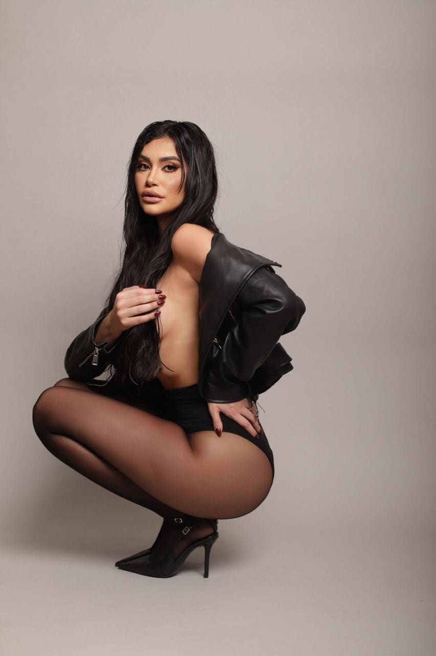 A woman crouches in a studio wearing a black leather jacket, high-waisted briefs, sheer tights, and pointed heels.
