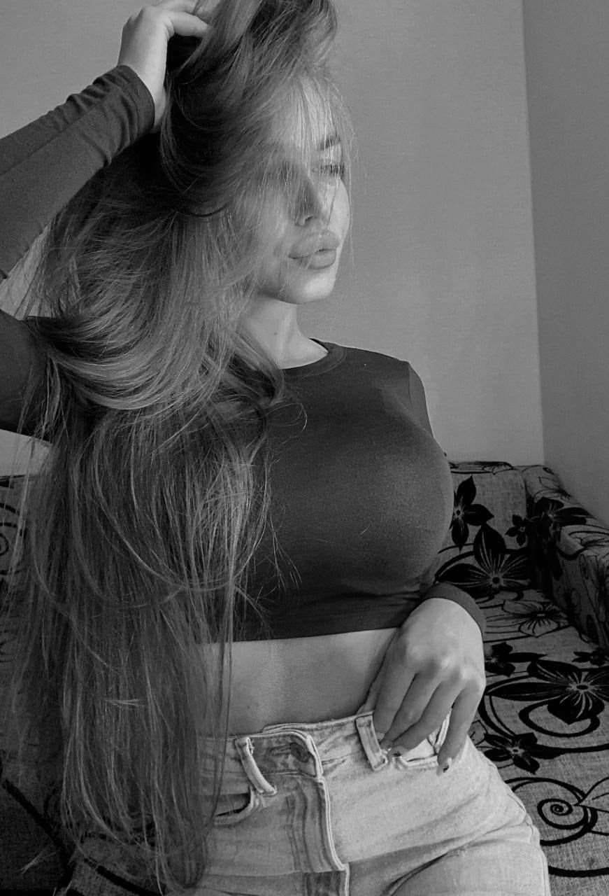 Seated on a floral couch, a woman in a dark crop top and light jeans rests one hand on her waist and touches her hair.