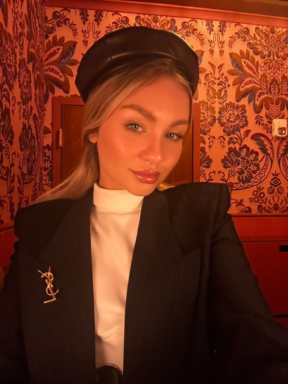 A woman in a black leather beret, white turtleneck, and pinstriped blazer poses before a floral patterned wall.