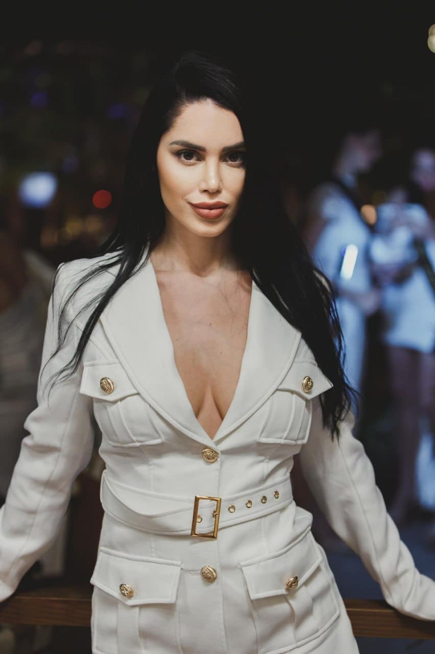 A woman in a white tailored jacket with a deep V-neck and gold buttons stands facing forward against a blurred indoor background with people.