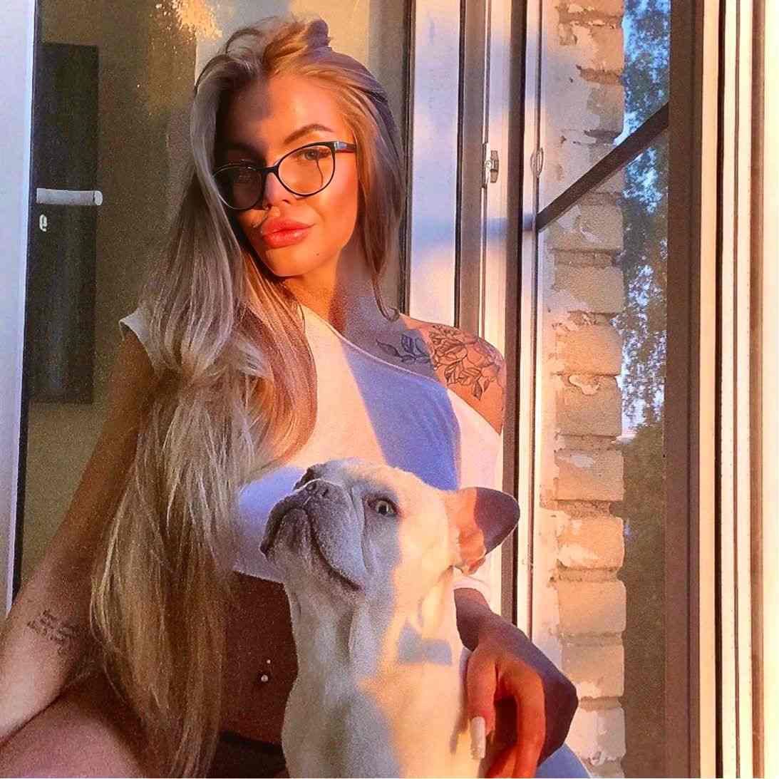 A woman with glasses holds a small white dog by a window, wearing a sleeveless top that reveals a shoulder tattoo.