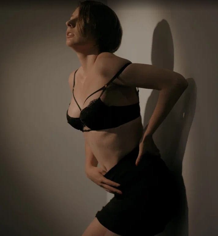 A woman in a black lace bra and shorts stands in profile with a hand on her hip, casting a strong shadow on the wall behind her.