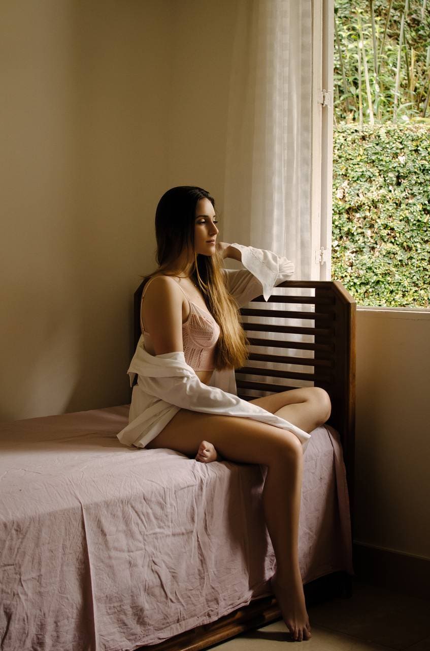 A woman sits on a bed with pink sheets, wearing a pink lace bra and draped white shirt, looking out a window.