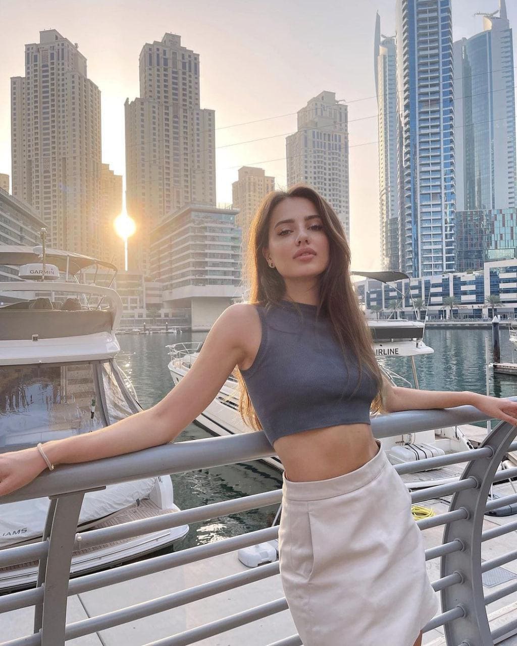 A woman leans on a waterfront railing at sunset, wearing a grey crop top and light skirt, with yachts and a city skyline behind her.